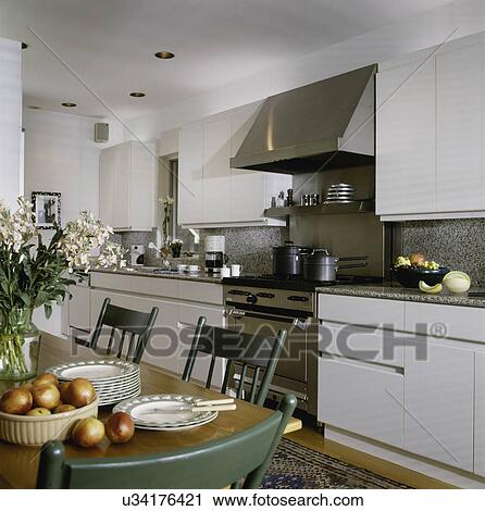 Stock Image - KITCHEN; with partial table showing to left with green chairs, and granite counters, stainless steel stove with white cabinets and sink. Fotosearch