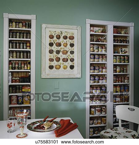 Stock Image - Kitchen with unique storage pantry set in wall filled with food items, light green wall, and framed print of cherries. Fotosearch