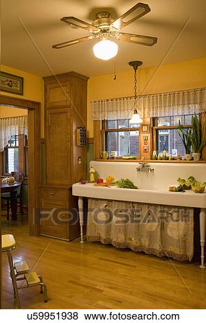 Stock Photo - KITCHENS: Arts and Crafts home, old fashioned long freestanding sink with embroidered vintage textile of swans, valance, window sill of corn collectibles, wood floor, built in cabinet next to sink. Fotosearch