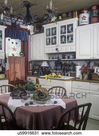 Stock Image - KITCHENS - Creatively decorated farmhouse kitchen. White cabinets, star motif used throughout. Tin chandelier decorated with twigs and berries. Table in foreground. Red accent color, tulips, rabbit,. Fotosearch