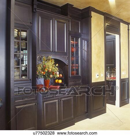 Stock Photo - KITCHENS - Detail of built in dish cabinet / buffet. Richly stained wood, alcove are with large bouquet of flowers, beige stone flooring. Doorway on right, yellow walls.. Fotosearch