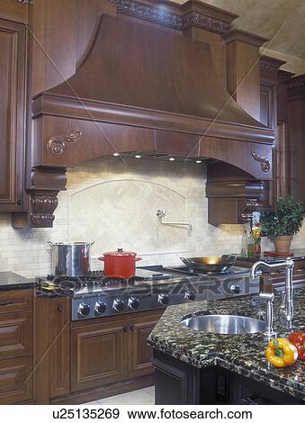 Stock Photo - KITCHENS - Large stove top area, grill, granite counters, rich wood cabinetry, high end, large elaborate wood exhaust hood, beige tile back splash, propped with colorful accents, red cooking pot, peppers,. Fotosearch