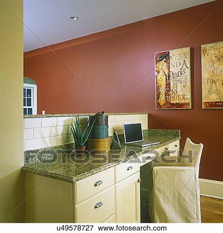 Stock Photo - KITCHENS - Open floor plan with kitchen on other side of 1/2 wall, cream wood built in desk with granite top. Laptop computer, slip covered chair, warm red wall.. Fotosearch