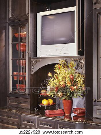 Stock Photo - KITCHENS - Richly stained wood built-in dish cabinet/buffet, door open to reveal TV. Below alcove with large bouquet of flowers, red pottery. Fotosearch