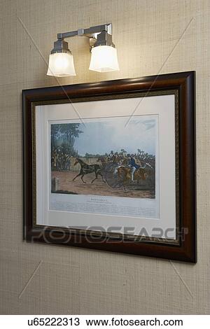 Stock Image - LIGHTING: An Arts and Crafts design influenced light sconce, a framed print of horse racing hangs under light, grass cloth wallpaper. Fotosearch
