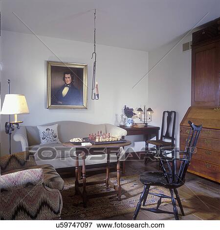 Stock Photo - LIVING ROOM - Camel back sofa. Antique furniture, Windsor chair, table with chess game, hanging candleholder, secretary, wide plank floors, white walls, simple plain Colonial, Early American. Fotosearch