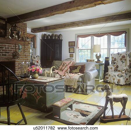 Stock Image - LIVING ROOM: country style, scarf drapes, grey leather sofa, draped quilt, painted trunk, painted checkerboard floor, horse on wheels, hooked rug,. Fotosearch