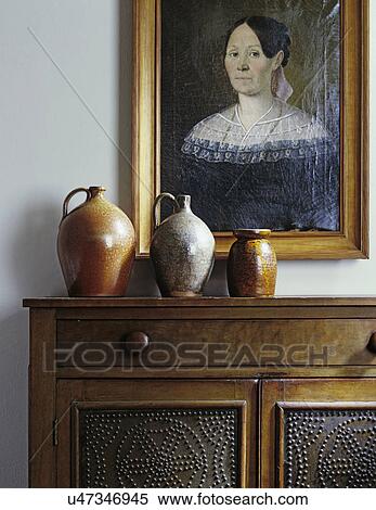 Stock Photography - LIVING ROOM DETAILS: Still life, sideboard with three salt glaze jugs, punched tin panels on walnut cabinet, portrait oil painting of woman, early American colonial style. . Fotosearch