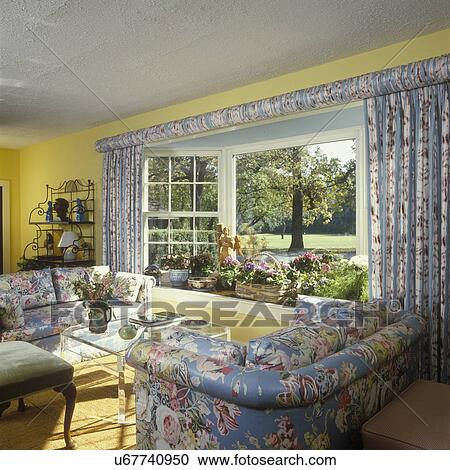 Stock Image - LIVING ROOM - Traditional Living Room - floral chintz upholstered matching sofas and drapery, gathered fabric on curtain rod, yellow walls, textured ceiling, plexiglass coffee table, metal baker's rack, baskets of flowering plants in bay window. Fotosearch