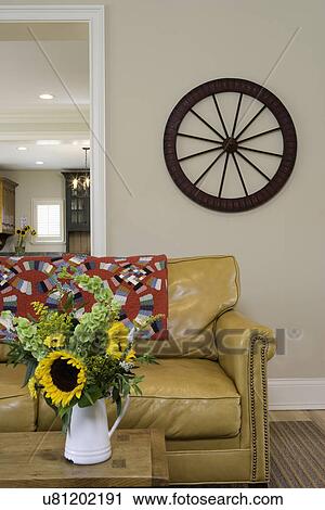 Stock Image - LIVING ROOMS: modern country, palomino colored leather sofa antique red quilt, enamel pot holds sunflowers and bels of Ireland, round game wheel on wall. Fotosearch