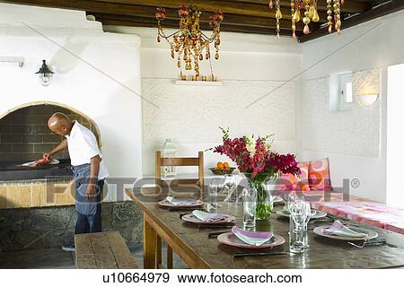 Stock Photo - Man cooking in dining room with wooden table in front, Mykonos, Greece. Fotosearch