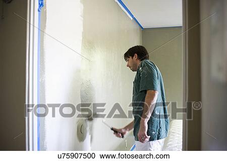 Stock Image - Man painting bedroom wall with paint roller. Fotosearch
