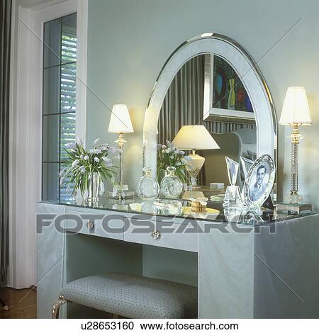 Stock Image - MASTER BEDROOMS - Sleek ladies vanity, arched mirror. Soft silver & aquamarine colors. Romantic, Art Deco and 1950's influence, crystal boudoir lamps, crystal perfume, silver frames, vase flowers . Fotosearch