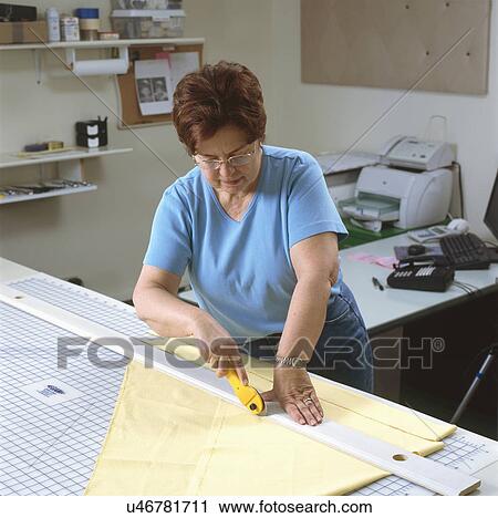 Stock Image - Mature woman cutting fabric in preparation for sewing on an electric sewing machine. Fotosearch
