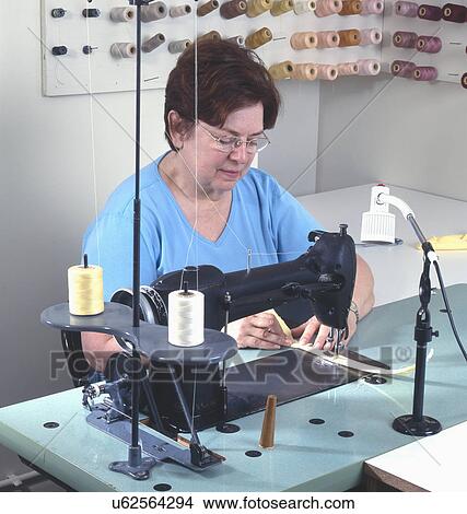 Picture - Mature woman using electric sewing machine with spools of thread stored on wall pegs in background. Fotosearch