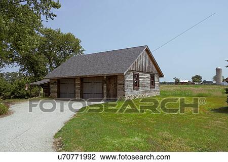 Stock Image - OUTBUILDINGS/ GARAGE: weathered rustic log garage, field stone foundation, two doors with a service door, two windows, lawn and driveway, horizontal, more sky showing with farm fields beyond and a silo. Fotosearch