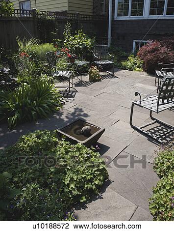 Stock Image - PATIO AREA: Fenced city garden and patio, small fountain in foreground, flower borders along wooden privacy fence. Fotosearch