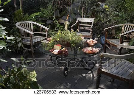 Stock Image - PATIO AREA: Four teak British colonial style furniture, glass top coffee table, container gardens cheese plate, tiki torch, potted plants. Fotosearch
