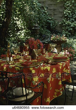 Stock Photography - PATIO AREA: Iron patio set, red tablecloth, gerberas, ivy and impatiens, lantern, gold and red table setting, close-up, wine bottle, wineglasses. Fotosearch