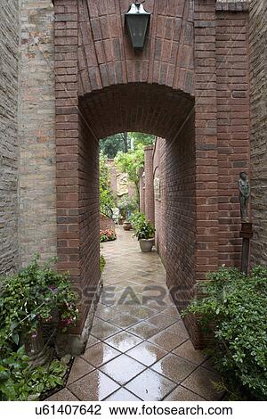 Stock Image - PATIO AREAS: Brick entry passage, lantern above, shade plants line walls, open courtyard beyond. Fotosearch