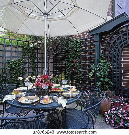 Stock Image - PATIOS: Close up on iron patio table with overhead umbrella, set for lunch, surrounded by trellis, city garden, pink impatiens. Fotosearch