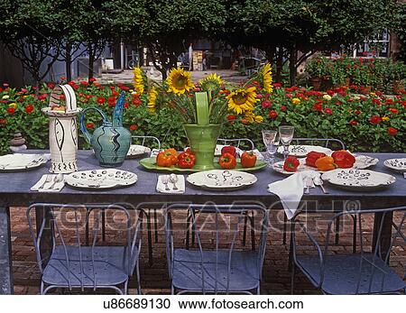 Stock Image - Patios: Detail of metal table set for dinner party, vase of sunflowers and red peppers create center piece, bed of red zinnias in background. . Fotosearch