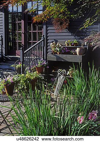 Stock Image - Patios: Red brick patio area, potting station on the right. Lots of potted flowers. Clapboard house. Metal table and chair.. Fotosearch