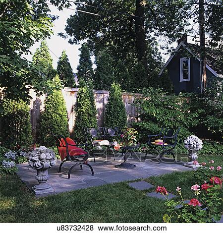 Stock Photo - PATIOS: Slate tile patio area along a privacy fence, black iron patio furniture, concrete sculpture urns, sun hat, red throw, geraniums in foreground.. Fotosearch