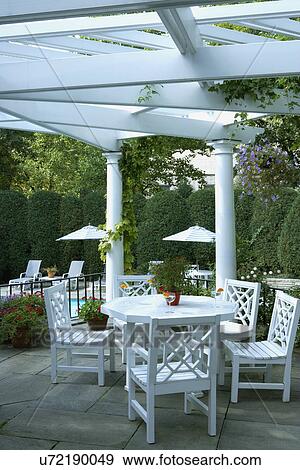 Stock Photo - PERGOLAS: Classical white columned pergola, grey slate patio, Chinese Chippendale style patio set, close up of dining area, overlooking pool, two patio umbrellas below at pool, lounge chairs in distance, black iron railing. Fotosearch