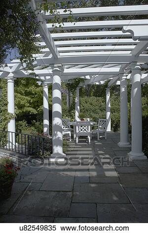 Stock Photography - PERGOLAS: Classical white columned pergola, grey slate patio, Chinese Chippendale style patio set, black iron railing to left straight on view, hanging plant in upper left corner of frame. Fotosearch