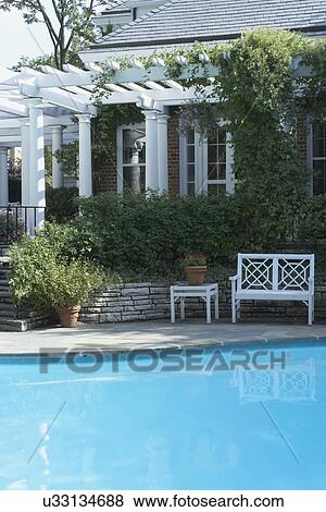 Stock Photo - PERGOLAS & POOLS: classical white columned pergola along a brick home, climbing vine, Chinese Chippendale style garden bench, swimming pool in foreground, closer version. Fotosearch