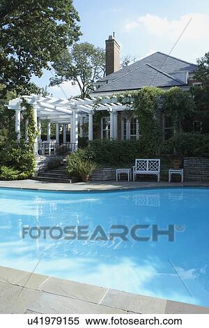 Stock Photography - PERGOLAS & POOLS: classical white columned pergola along a brick home, climbing vine, Chinese Chippendale style garden bench, swimming pool in foreground, closer version. Fotosearch