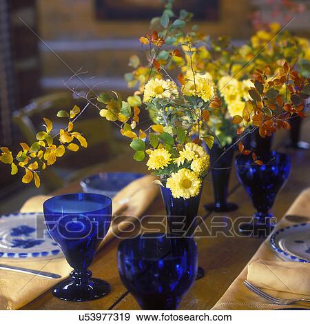 Stock Photo - PLACE SETTINGS: Plank table top, blue goblets yellow, flowers, orange leaves, yellow place mats, napkins, fall, harvest, thanksgiving . Fotosearch