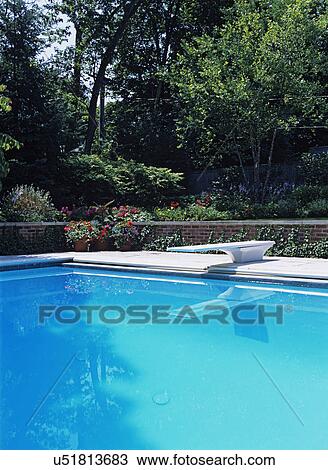 Stock Image - Pool: Close up of deep end of 20' x 46' lap pool, diving board, nice shot of the elevated landscaped lawn and garden, brick retaining wall, potted flowers, trees in background.. Fotosearch