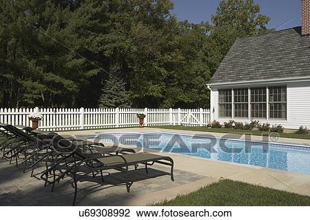 Stock Image - Pools: View towards house, four windows, white picket fence on the left, chaise lounges foreground. Partial view of rectangular pool with half circles at each end. Trees in background.. Fotosearch
