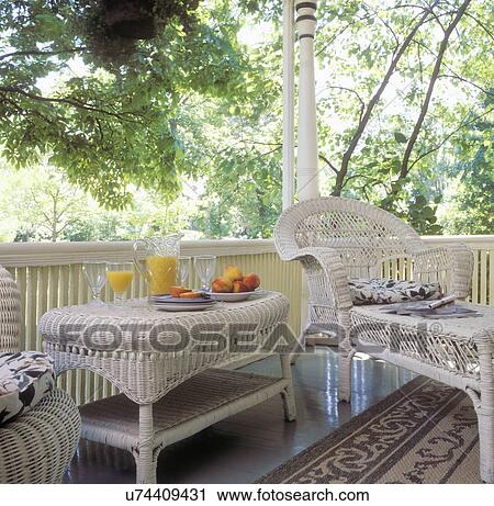 PORCHES: Detail of white wicker furniture, a side table with juice and peaches, chaise lounge with magazine and cushions, white railing, painted wood floor, area rug, trees in background View Large Photo Image Stock Image - PORCHES: Detail of white wicker furniture, a side table with juice and peaches, chaise lounge with magazine and cushions, white railing, painted wood floor, area rug, trees in background. Fotosearch