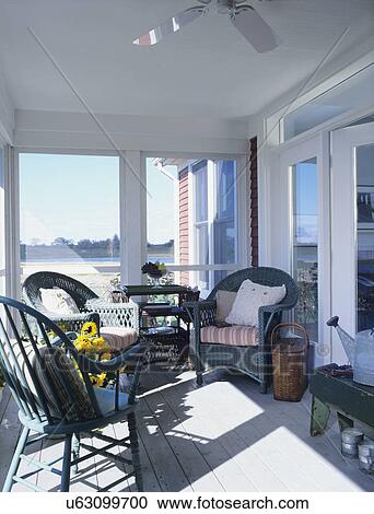 Stock Image - PORCHES: Screened porch overlooking lake, green wicker chairs with striped cushions, low back Windsor painted green, sunflowers, angled green bench with watering cans, new built farmhouse, ceiling fan, transom over doorway. Fotosearch