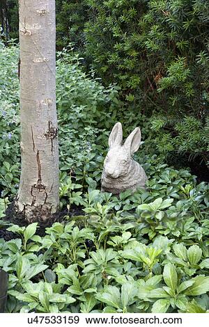 Stock Photo - Rabbit sculpture by tree trunk surrounded by plants in park. Fotosearch