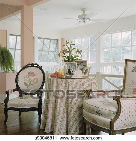 SITTING AREAS: Two chairs with floral fabric, table with striped cloth, family photos, column to left with fern plant windows behind View Large Photo Image Stock Image - SITTING AREAS: Two chairs with floral fabric, table with striped cloth, family photos, column to left with fern plant windows behind. Fotosearch