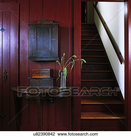 Stock Image - Stairways: Bottom of enclosed staircase, restored 1841 home, wood panels painted a deep red, antique half circle side table with wooden boxes on left. . Fotosearch