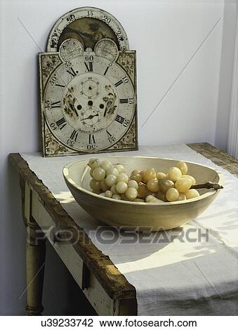 Stock Image - STILL LIFE: worn vintage farm table with antique clock face and wooden bowl full of stone grapes. Fotosearch