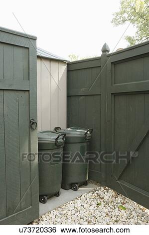 Stock Photograph - STORAGE: Dark green painted privacy fence, garbage bins behind gate on a concrete slab, gravel. Fotosearch