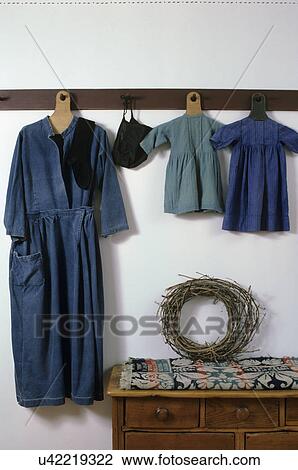 Stock Image - TEXTILES: An Amish maternity dress and antique children's clothing hang on a peg rack, 1844 coverlet folded on dresser below, grapevine wreath. Fotosearch