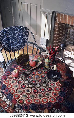 Stock Image - TEXTILES: hand woven Penny rugs on display on round table and Windsor arm chair, Jar of licorice, toleware, colonial sage green painted fireplace behind. Fotosearch