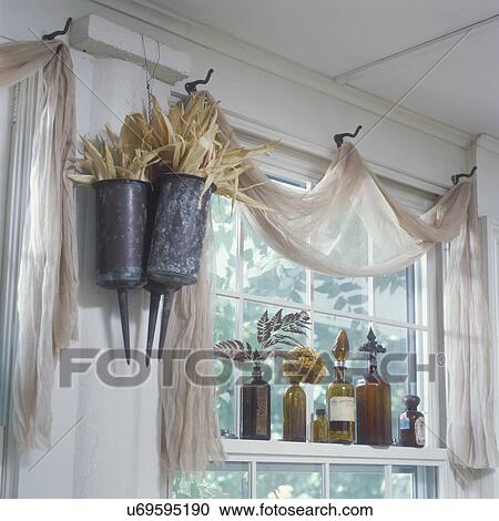 Stock Image - WINDOW TREATMENTS: Unique, Scrim (a net like fabric) valance, old coat hooks serve as drape points for scarf valance, copper funeral urns on left filled with cornhusk, brown colored bottles on window sill . Fotosearch