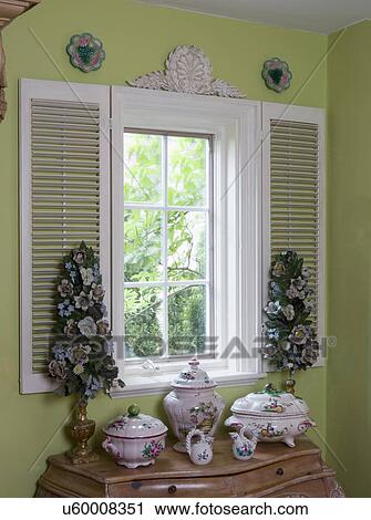 Stock Image - WINDOW TREATMENTS: White stationary shutters are decorative accents around window, architectural accent above, Faience pottery, metal enamel flower trees, lime green painted walls. Fotosearch