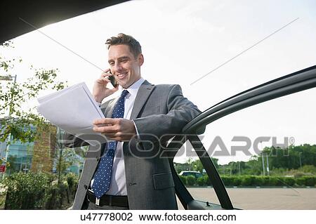 Stock Image - Businessman leaning against car door reading paperwork. Fotosearch