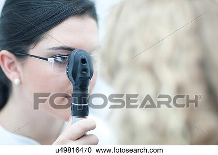 Stock Image - Female optician looking through ophthalmoscope. Fotosearch