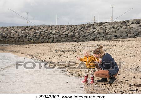 Stock Image - Mother and toddler crouching at waters edge. Fotosearch