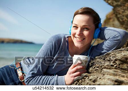 Stock Image - Portrait of young woman at coast with coffee and earphones. Fotosearch